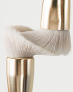 Close-up of two Seint makeup brushes with soft white bristle heads and gold ferrules pressed together