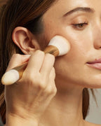 Hand blends cream makeup onto a model's cheek with a double-ended gold Seint brush