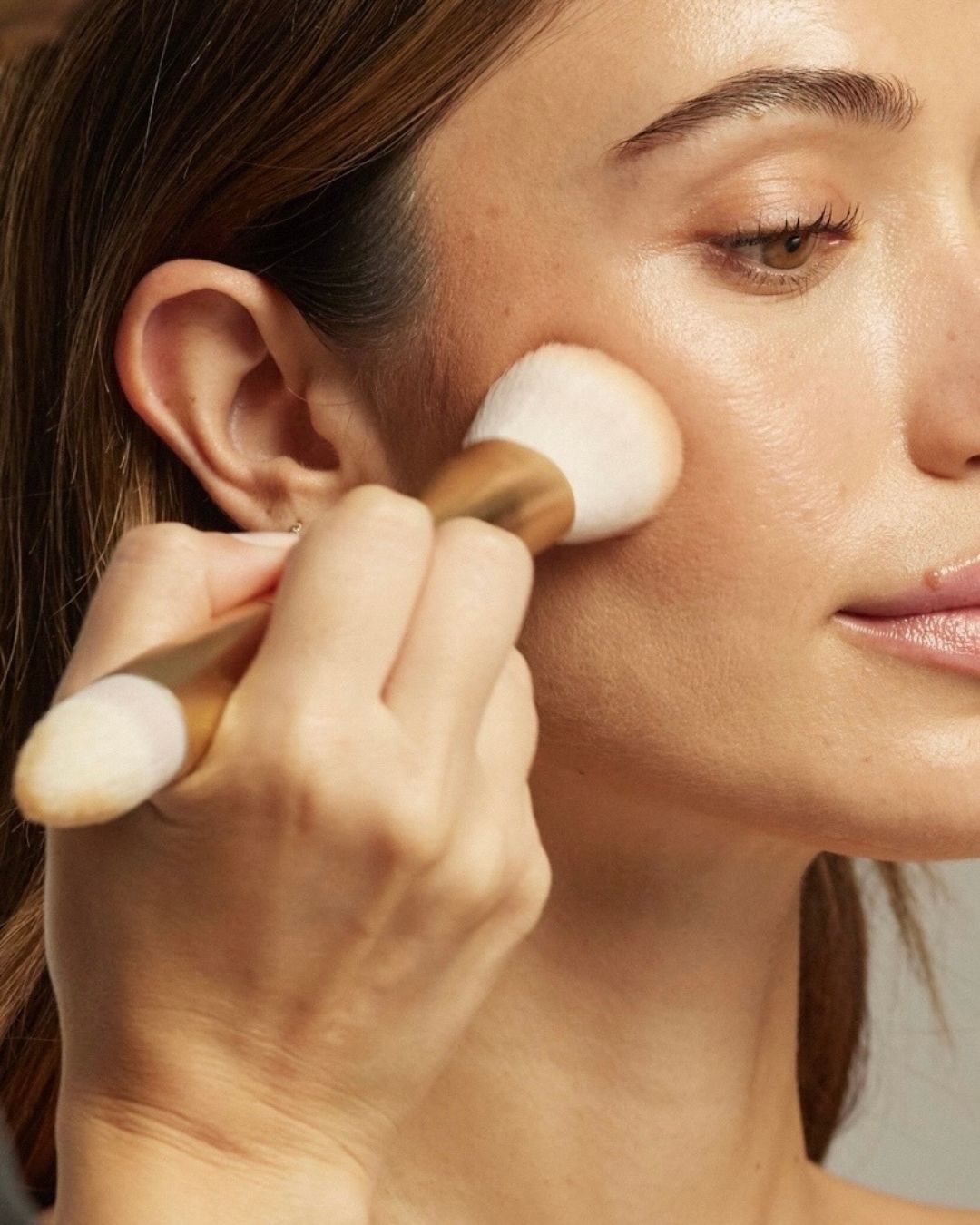 Hand blends cream makeup onto a model's cheek with a double-ended gold Seint brush
