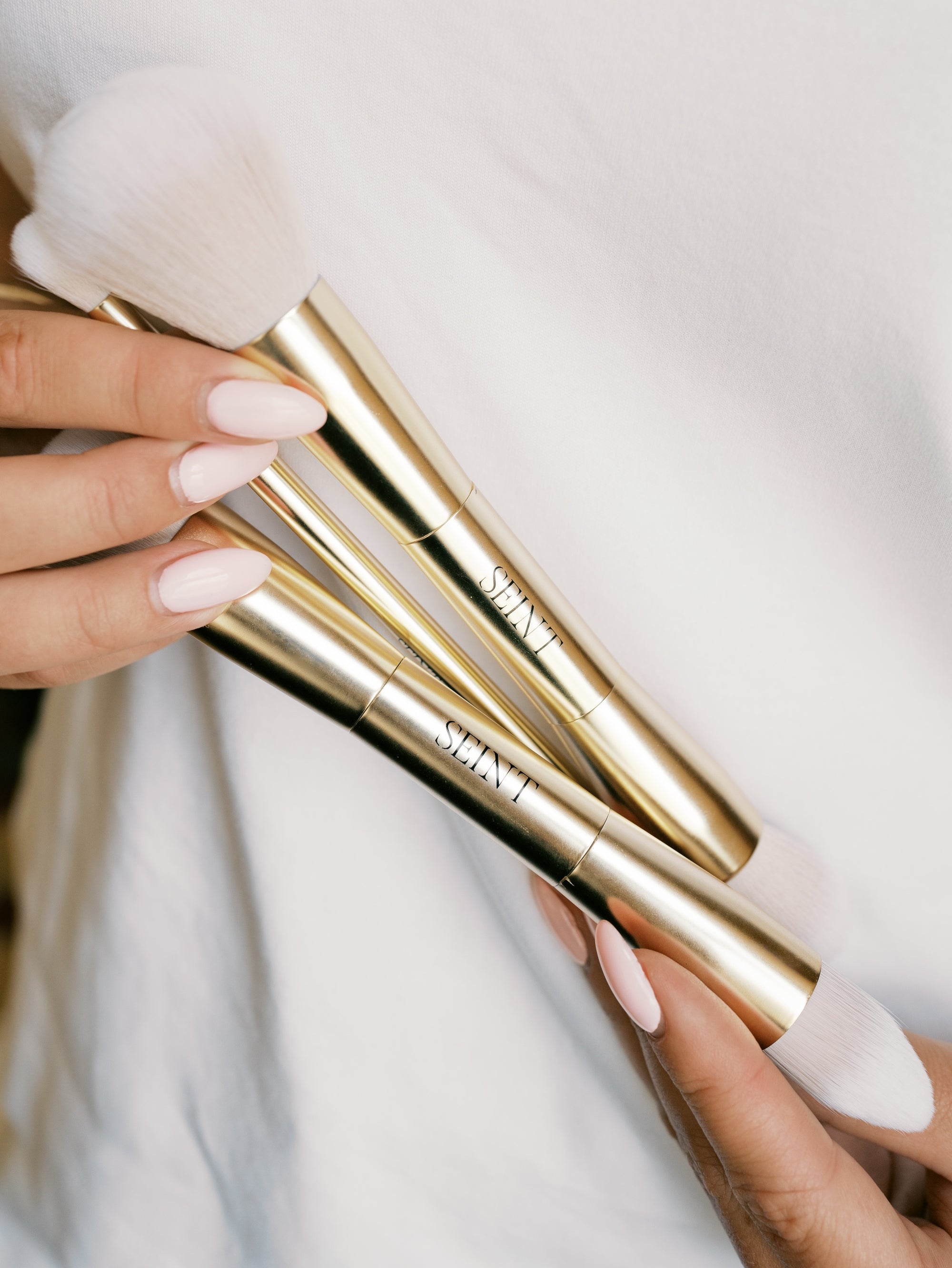 Trio of Seint makeup brushes held in hands, gold handles with SEINT wordmark and soft white bristle ends