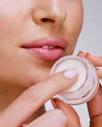 Seint Lip Balm jar held near a model's chin as she dips a fingertip into the creamy ivory balm