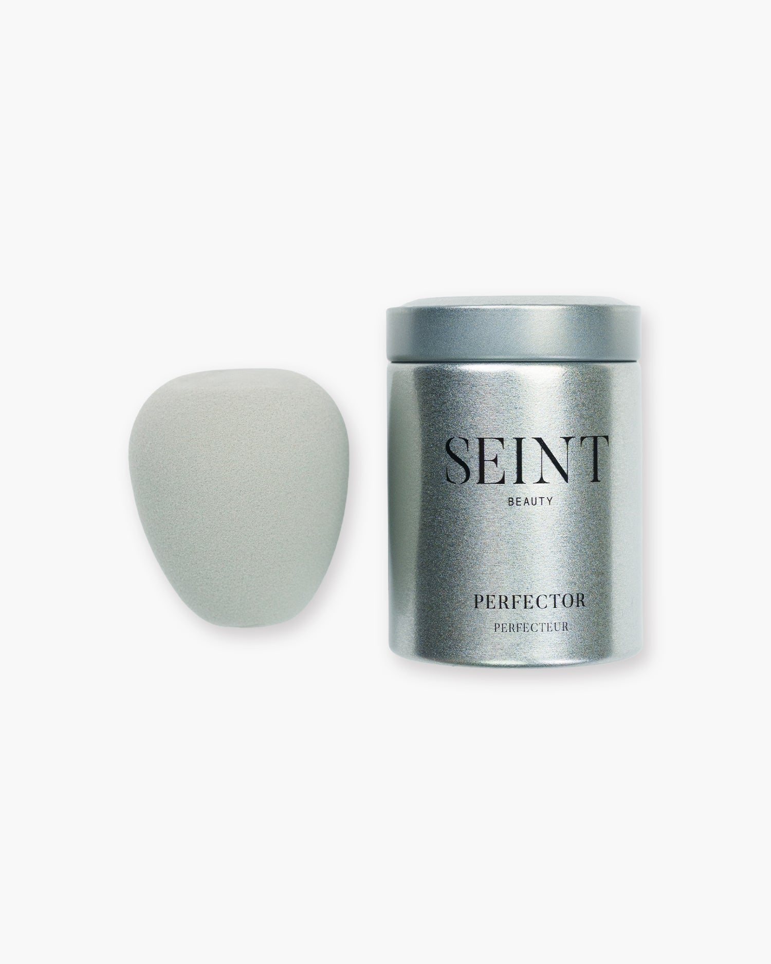 Seint Perfector blending sponge in soft gray beside its closed silver storage tin with Seint Beauty label