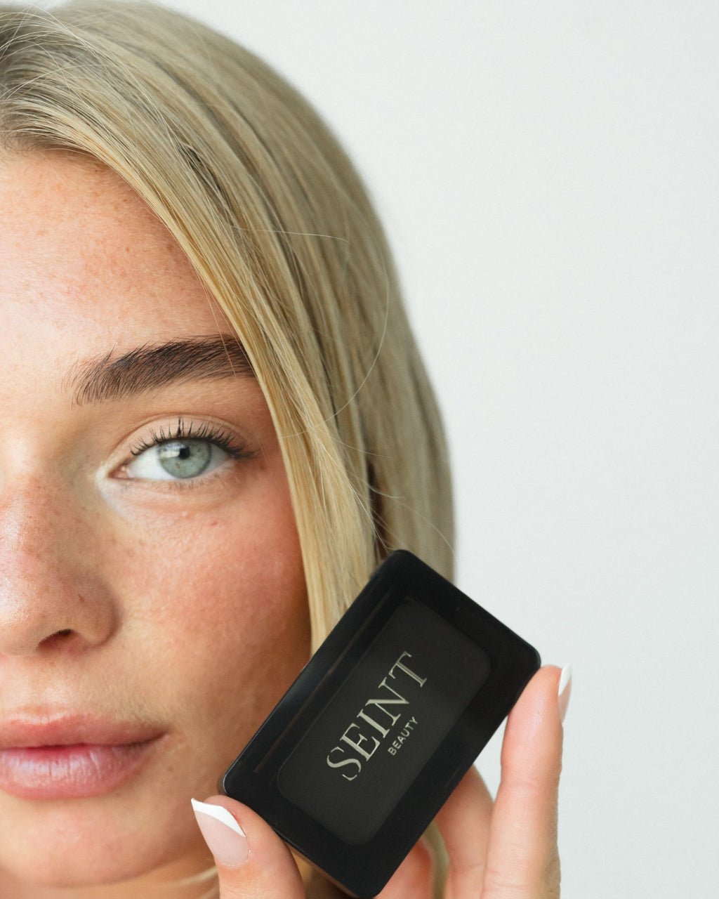 Seint Mini Compact held closed beside a model's cheek, its gold SEINT logo showing the compact's palm-size scale