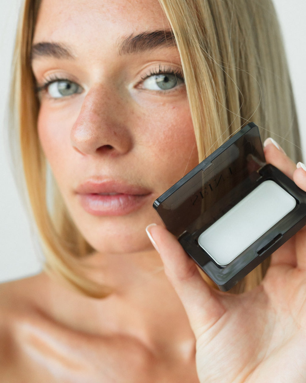 Seint Mini Compact open in a model's hand, revealing a white balm tin as she holds it near her face