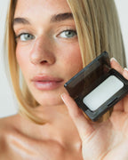 Seint Mini Compact open in a model's hand, revealing a white balm tin as she holds it near her face