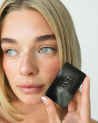 Seint Mini Compact closed at a model's fingertips as she glances aside, gold SEINT Beauty logo on the lid