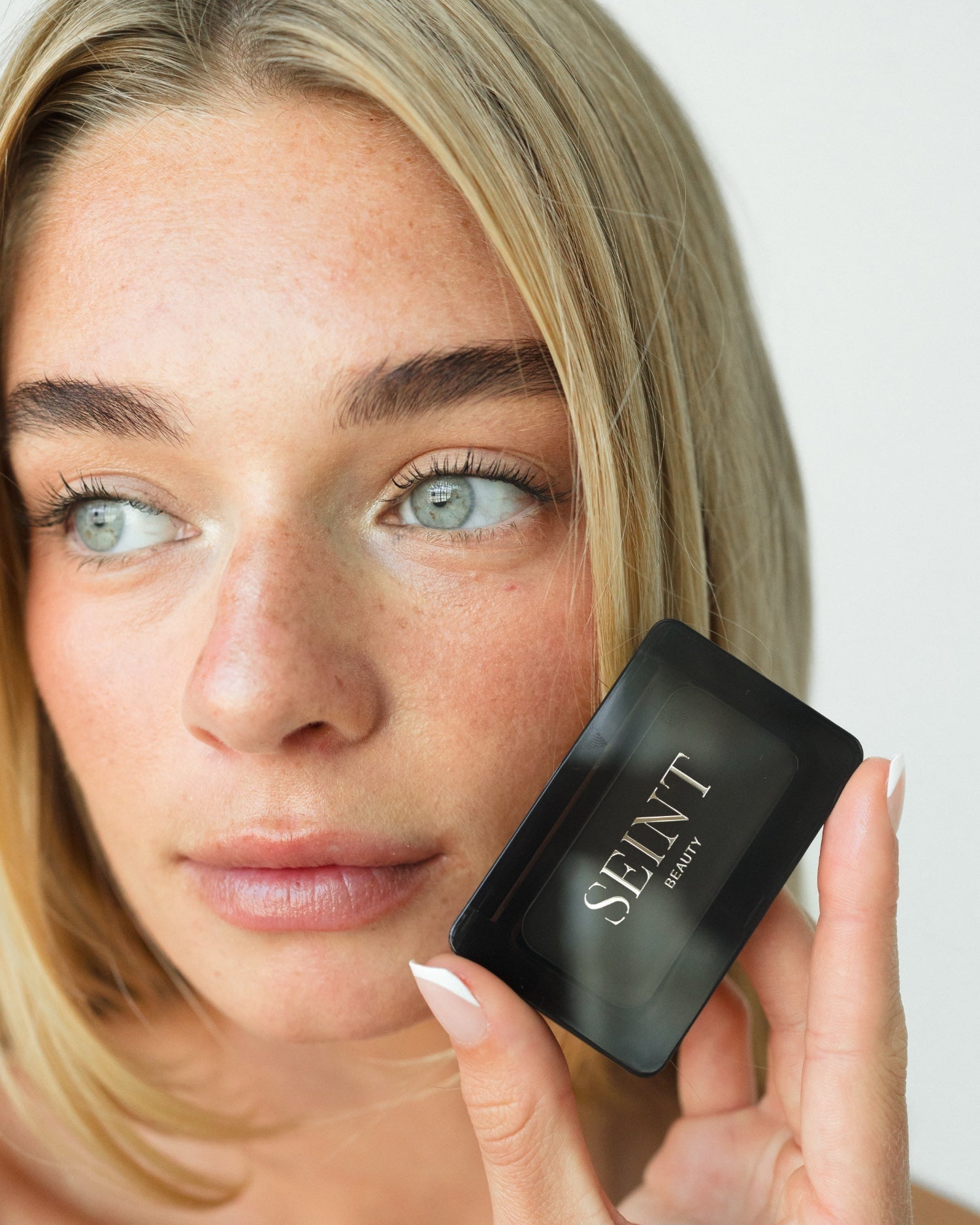 Seint Mini Compact closed at a model's fingertips as she glances aside, gold SEINT Beauty logo on the lid