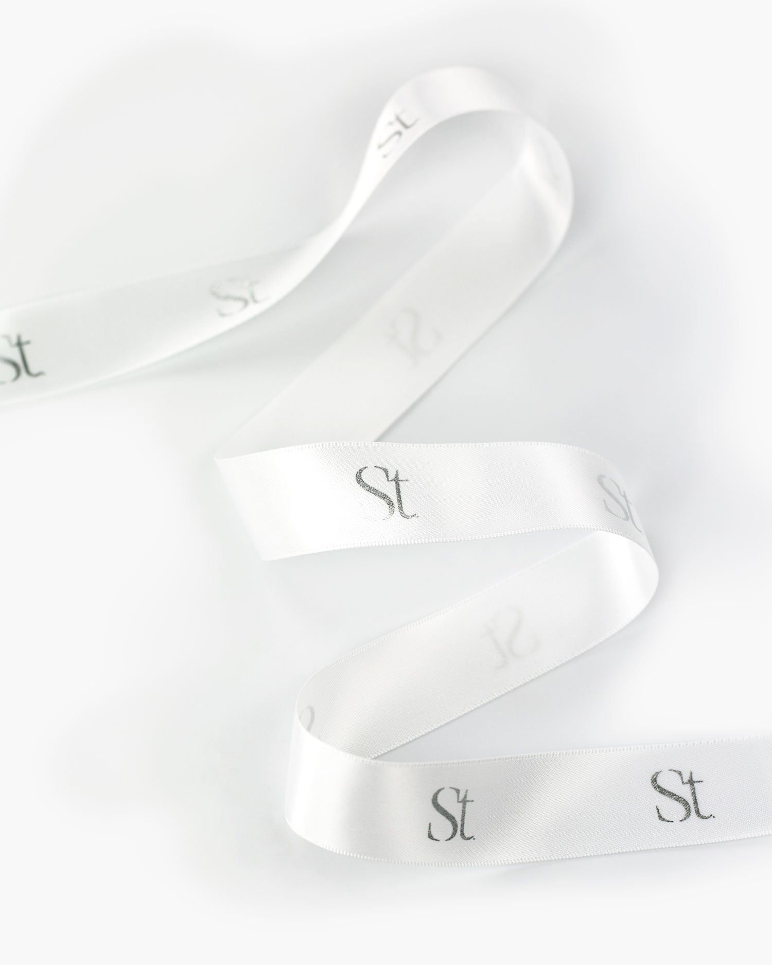 Seint Monogram Ribbon in Ivory: white satin ribbon looping softly, printed with the gray Seint monogram repeat