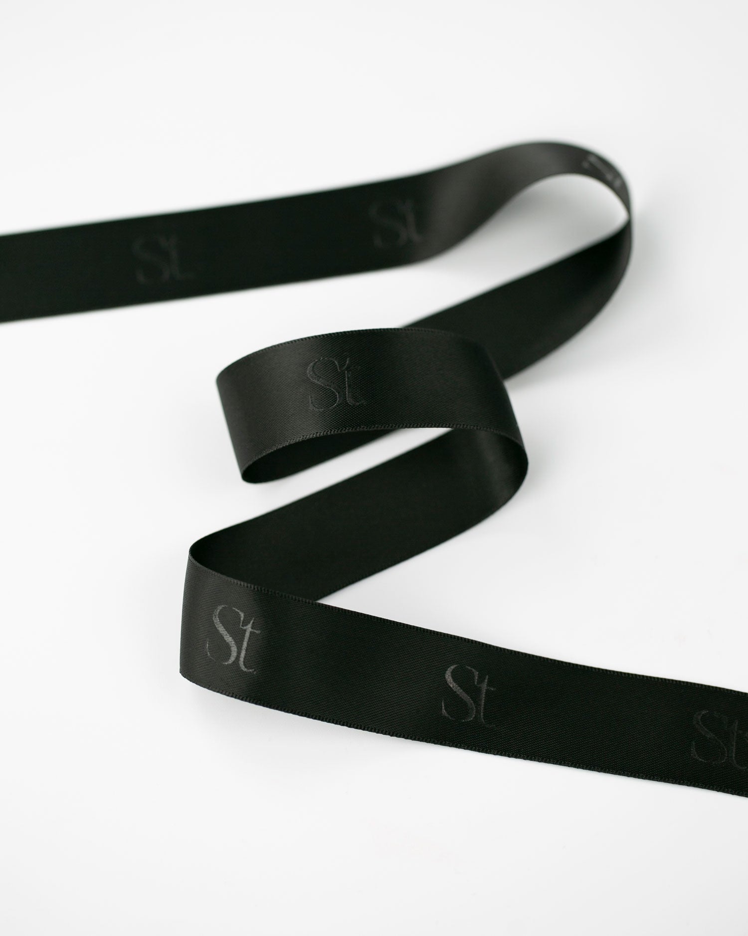 Seint Monogram Ribbon in Onyx, black satin curling loosely with the repeating tone-on-tone Seint monogram