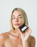 Seint Saved Bundle held open beside a model's face, white salve visible in the black mini compact